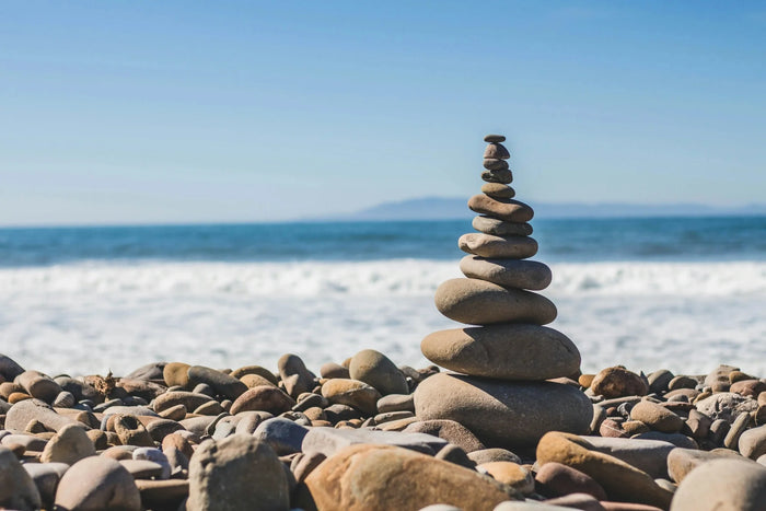 A stack of smooth stones on a rocky beach with waves in the background and a clear blue sky above.