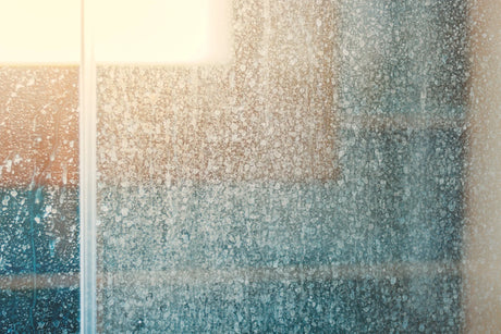 Close-up view of a foggy glass window with water droplets and condensation, partially illuminated by sunlight in the upper left corner.