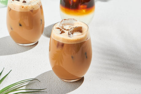Three glasses of iced coffee with frothy tops sit on a white surface, casting soft shadows.