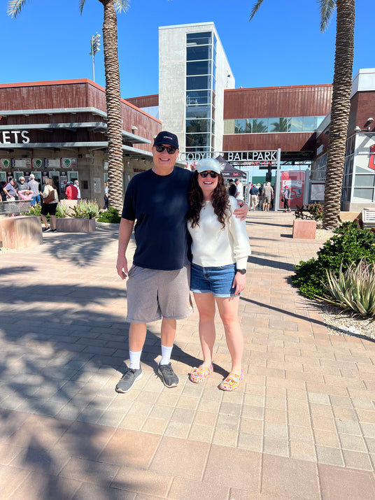 A man and woman stand smiling in front of a ballpark entrance on a sunny day.