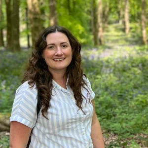 Woman with curly hair in a striped shirt smiles in a forest setting. Lush greenery and sunlight in the background.