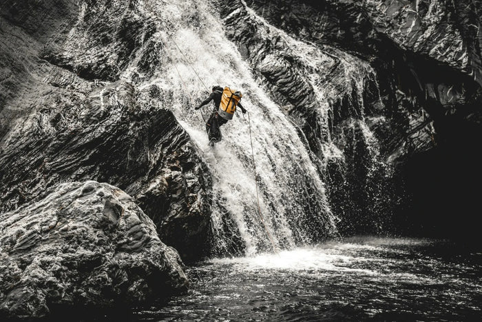 Person wearing a yellow backpack climbs a waterfall in a rocky landscape.