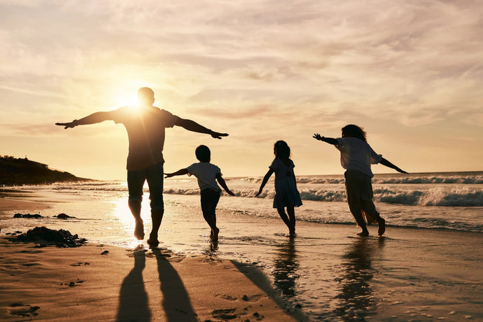 A group of four people, two adults, and two children, walk along the beach at sunset with their arms outstretched, casting long shadows on the sand.