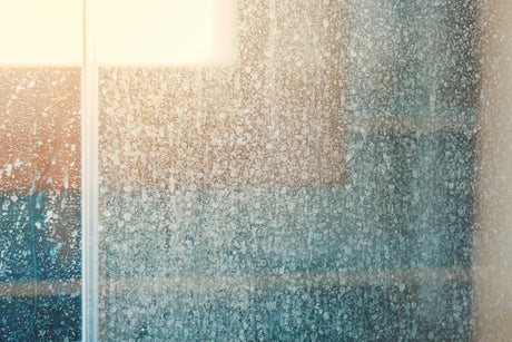 Close-up view of a foggy glass window with water droplets and condensation, partially illuminated by sunlight in the upper left corner.