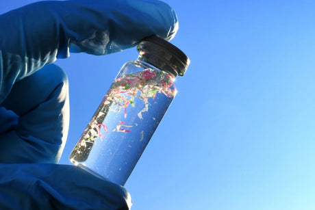 A gloved hand holds a clear vial containing small plastic particles against a bright blue sky.