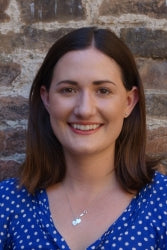 A woman with shoulder-length brown hair wearing a blue polka-dot blouse and a necklace, smiling in front of a brick wall.